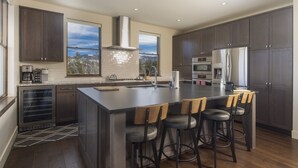 A modern kitchen with dark wood cabinets, a large island with bar stools, stainless steel appliances, and a view of the city through large windows. - Modern Elegance, Stunning Mountain Views, Hot Tub, Fire Pit, Walk to River! (Breckenridge)