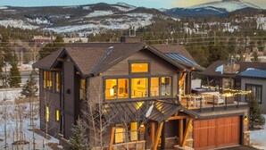 A modern two-story house with a large deck and a garage, surrounded by a snowy landscape and mountains. - Modern Elegance, Stunning Mountain Views, Hot Tub, Fire Pit, Walk to River! (Breckenridge)