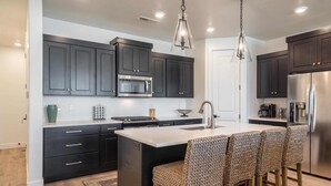 A modern kitchen with dark cabinets, a central island with bar stools, stainless steel appliances, and pendant lighting. - Desert Azul by RedAwning (St. George)