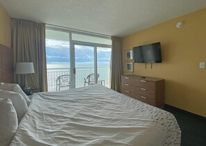 A hotel room with a bed, a TV mounted on the wall, a dresser, and a balcony with a view of the sea. - Camelot by the Sea by Palmetto Vacations (Myrtle Beach)