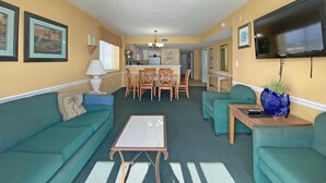 A compact living space with a dining area, kitchenette, and a flat-screen TV mounted on the wall. - Camelot by the Sea by Palmetto Vacations (Myrtle Beach)