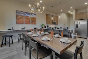 A modern kitchen with a wooden dining table set for six, stainless steel appliances, and a chandelier.