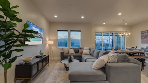 A spacious living area with a gray sectional sofa, a black console table, and a large window offering a view of a house and mountains. - Snow Canyon Oasis, Ocotillo 42, Large Private Pool and Hot Tub, 2 Firepits, 2 PlayStations (Santa Clara)