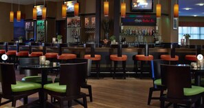 A restaurant with wooden flooring, red and green upholstered chairs, and a bar area with a minibar and bottles.