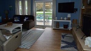 A living room with a sofa, coffee table, and a TV. There is a fireplace and a view of the outdoors through a glass door.