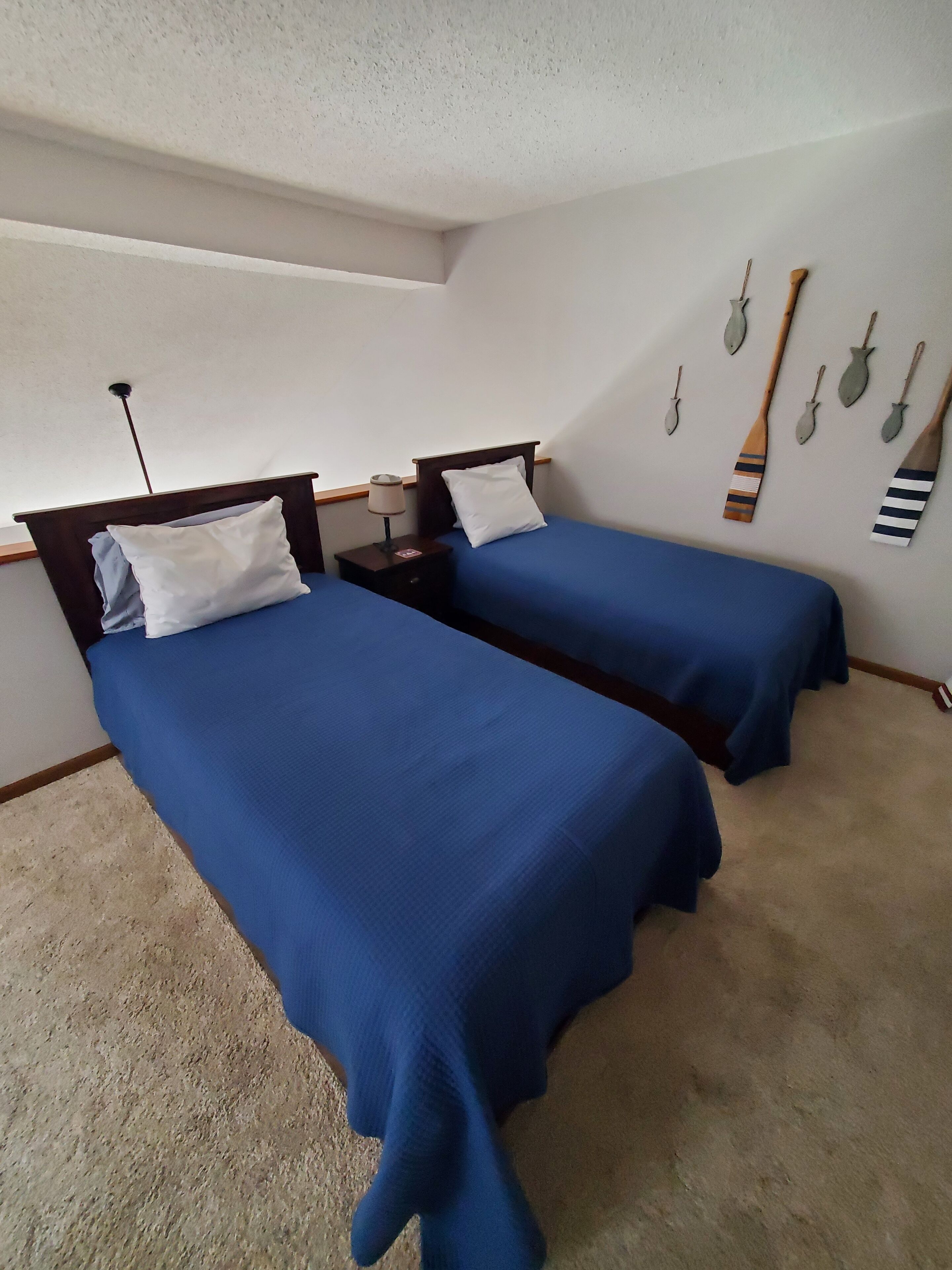 Una habitación con dos camas, cada una con cubrecama azul y almohadas blancas, una mesita de noche con una lámpara, y una pared decorada con objetos que parecen remos.