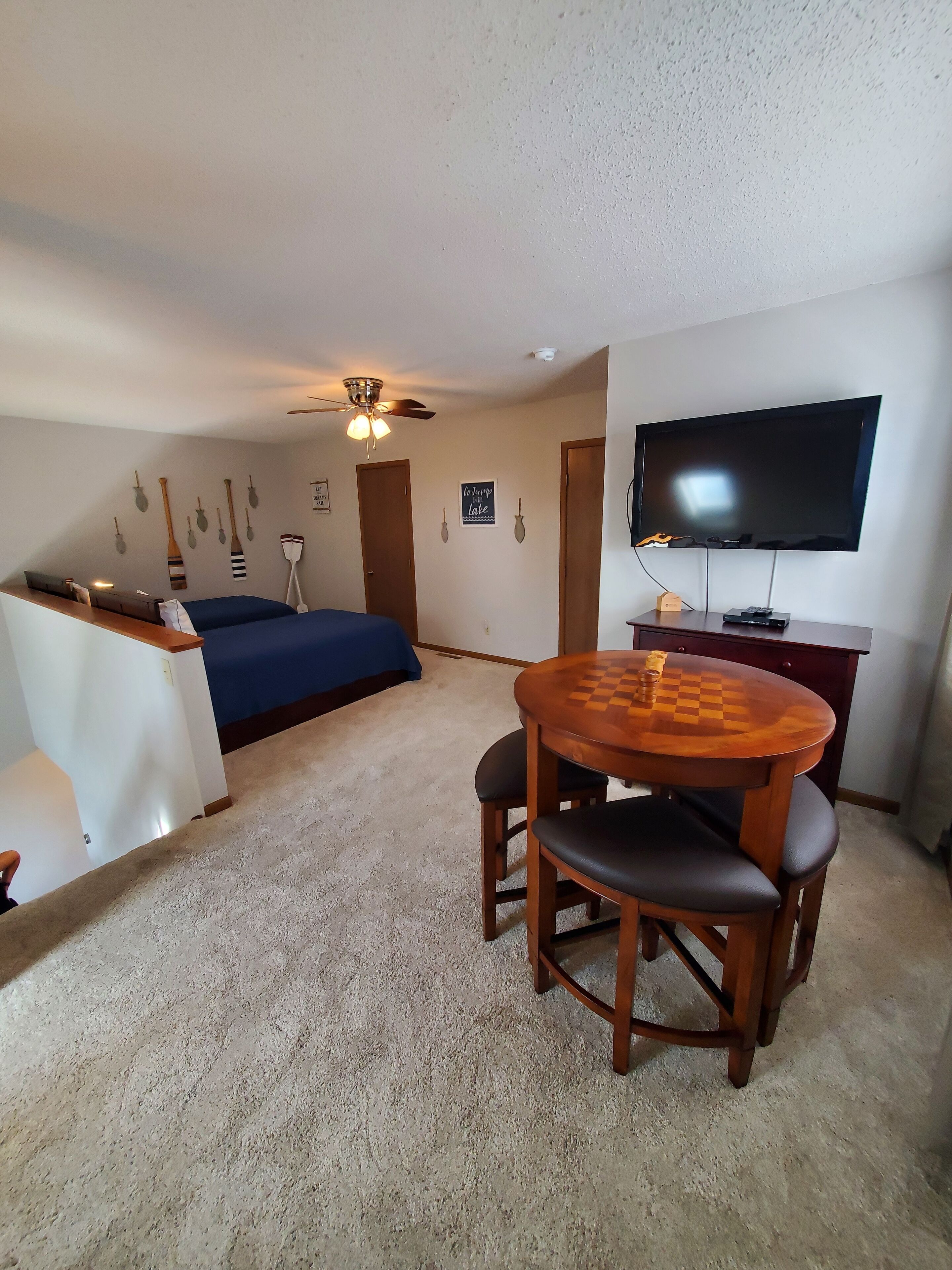 Ein Zimmer mit Bett, Schreibtisch mit Fernseher, Deckenventilator und Holztisch mit Stühlen.