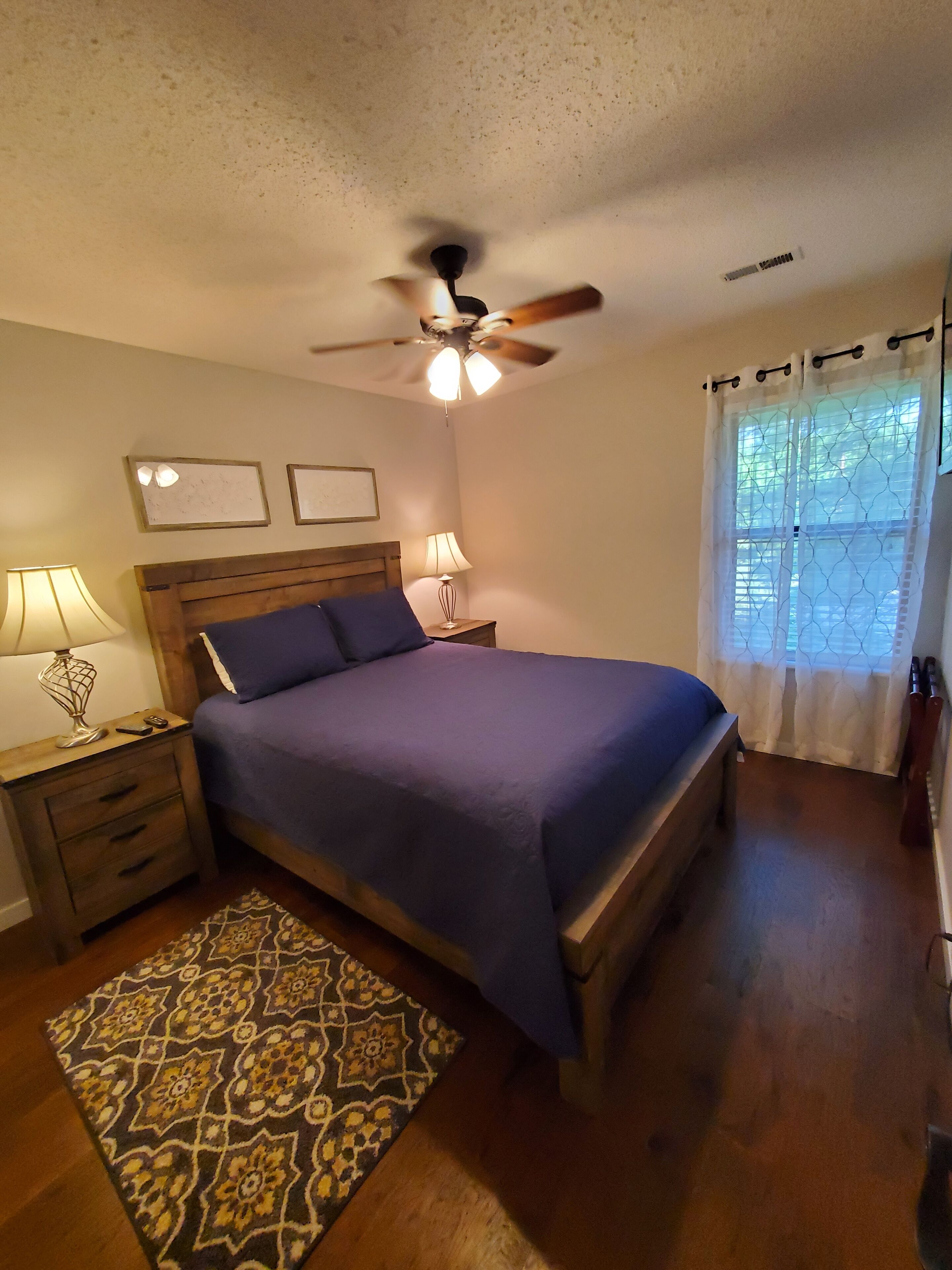 A bedroom with a bed, nightstands, a ceiling fan, a patterned rug, and a window with curtains.