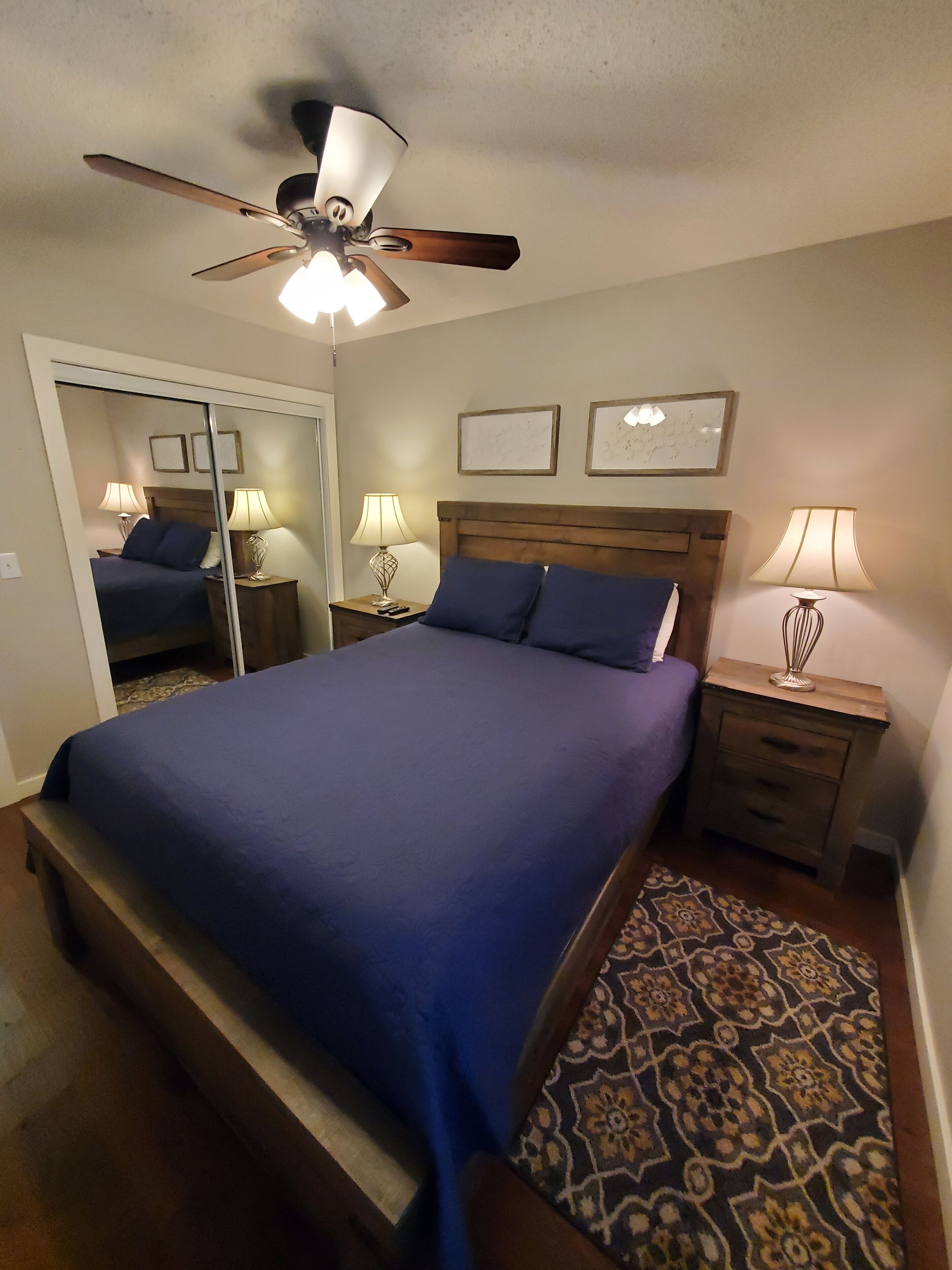 A bedroom with a bed, a ceiling fan, two bedside tables with lamps, a patterned rug, and a mirror.