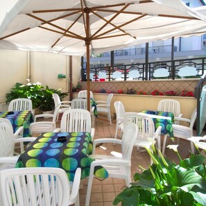 Een terras buiten met witte plastic stoelen en tafels, sommige met kleurrijke tafelkleden, onder een grote parasol.