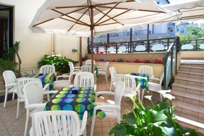 Een terras buiten met witte plastic stoelen en tafels, waarvan sommigen versierd zijn met kleurrijke tafelkleden, onder een grote parasol.