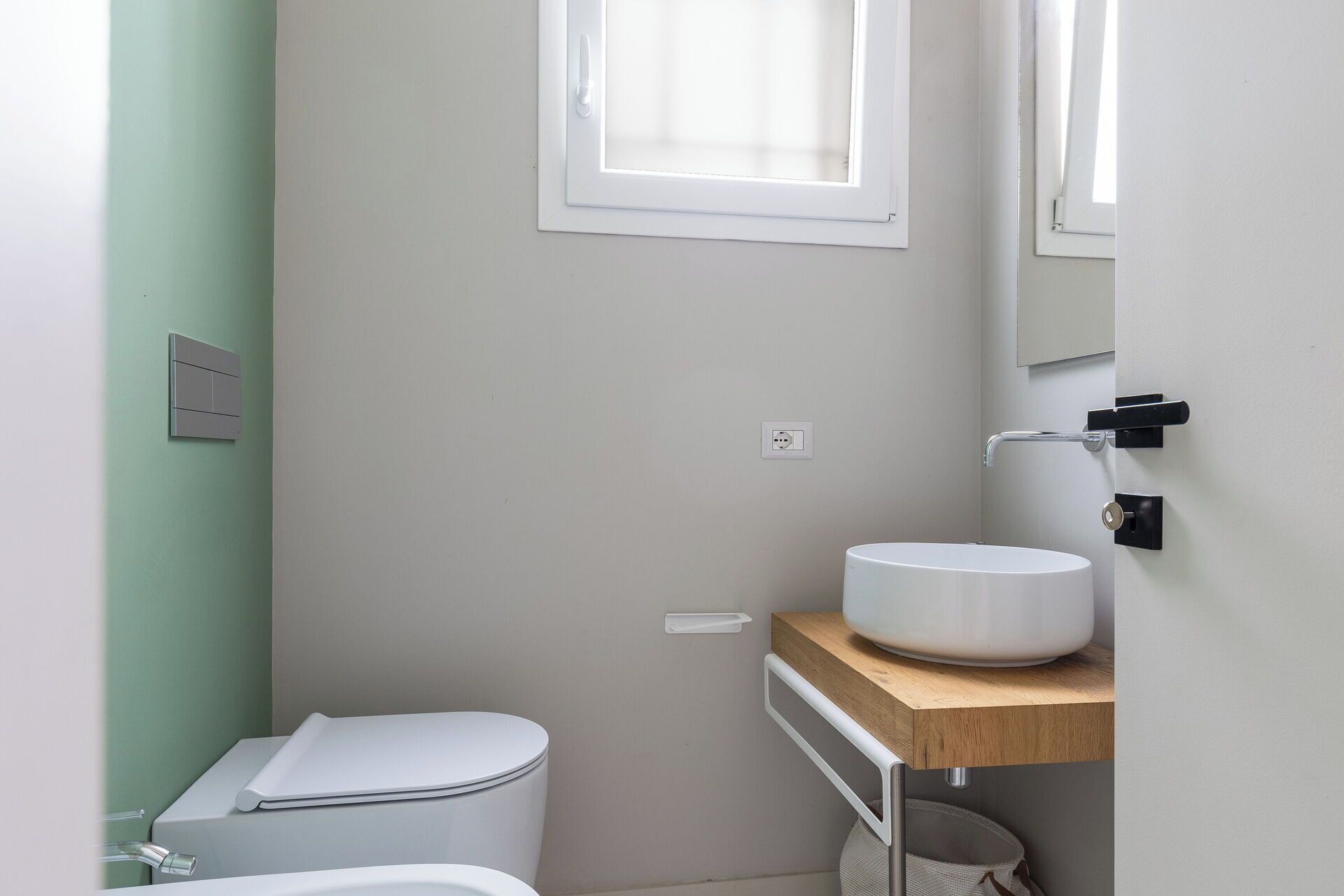 Ein kleines, modernes Badezimmer mit Toilette, einem Holzwaschtisch und einem weißen Waschbecken sowie einem Fenster über der Toilette.