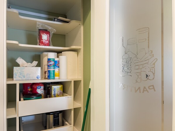 A kitchen with shelves stocked with cleaning supplies, a coffee maker, and a decorative wall with a cartoon character. - The Sunset House | Pool, Hot Tub & Putting Green (Tempe)