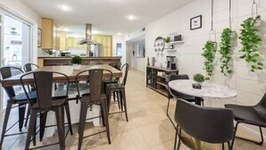 Ett modernt kök med en central köksö, en matplats med stolar och en bardisk med växter och kaffebryggare.