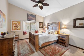 Une chambre à coucher avec un lit en bois, un ventilateur de plafond, une table de chevet avec une lampe, une commode et deux tapis.