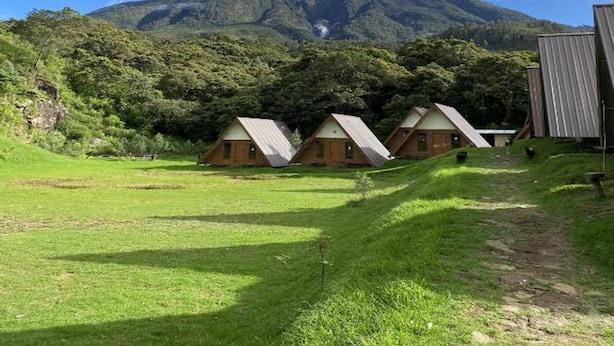 A row of wooden cabins with pitched roofs, set against a backdrop of a lush green landscape and a mountain with clouds. - Supercamp Sakura Hills (Tawangmangu)