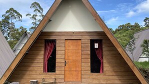 A triangular wooden cabin with a central door and two windows, surrounded by a grassy area and rocks. - Supercamp Sakura Hills (Tawangmangu)