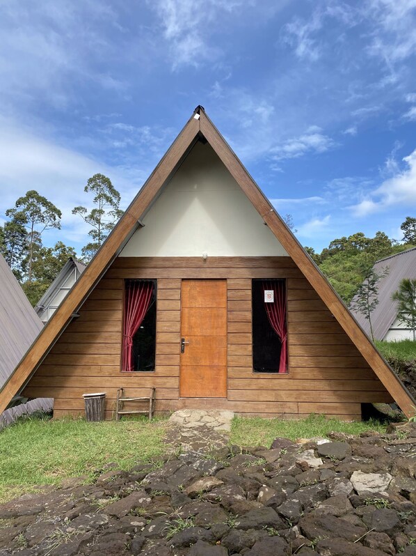 A triangular wooden cabin with a central door and two windows, surrounded by a grassy area and rocks. - Supercamp Sakura Hills (Tawangmangu)