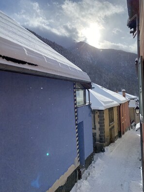 A narrow snow-covered street with buildings on either side, including a blue building with a window and a brown building with a chimney.