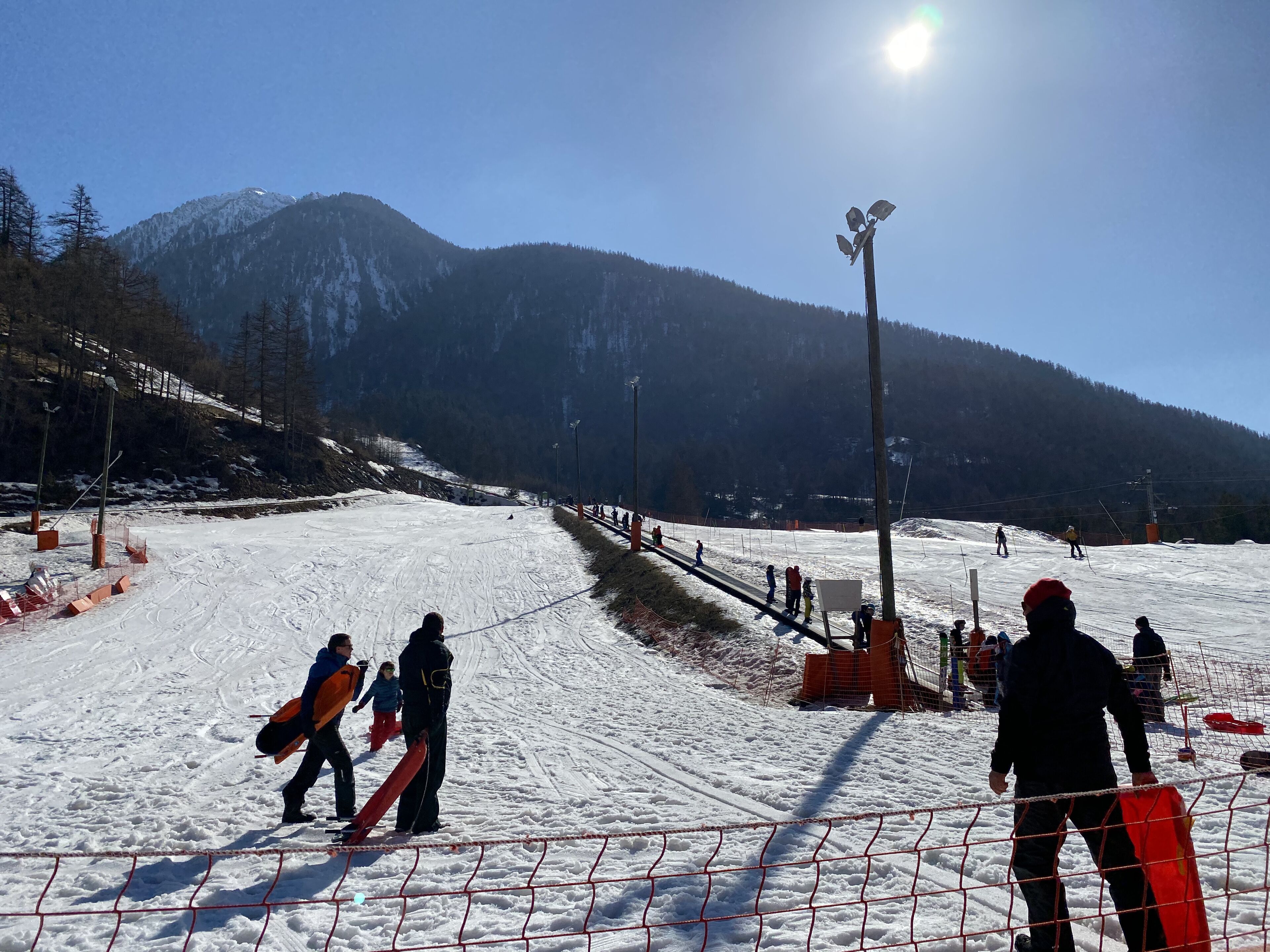 A snowy ski slope with a mountain in the background, a fence, and people with snowboards.