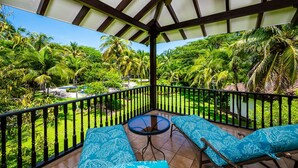 A covered balcony with two blue patterned sofas and a glass table, overlooking a lush garden with palm trees and a pool.