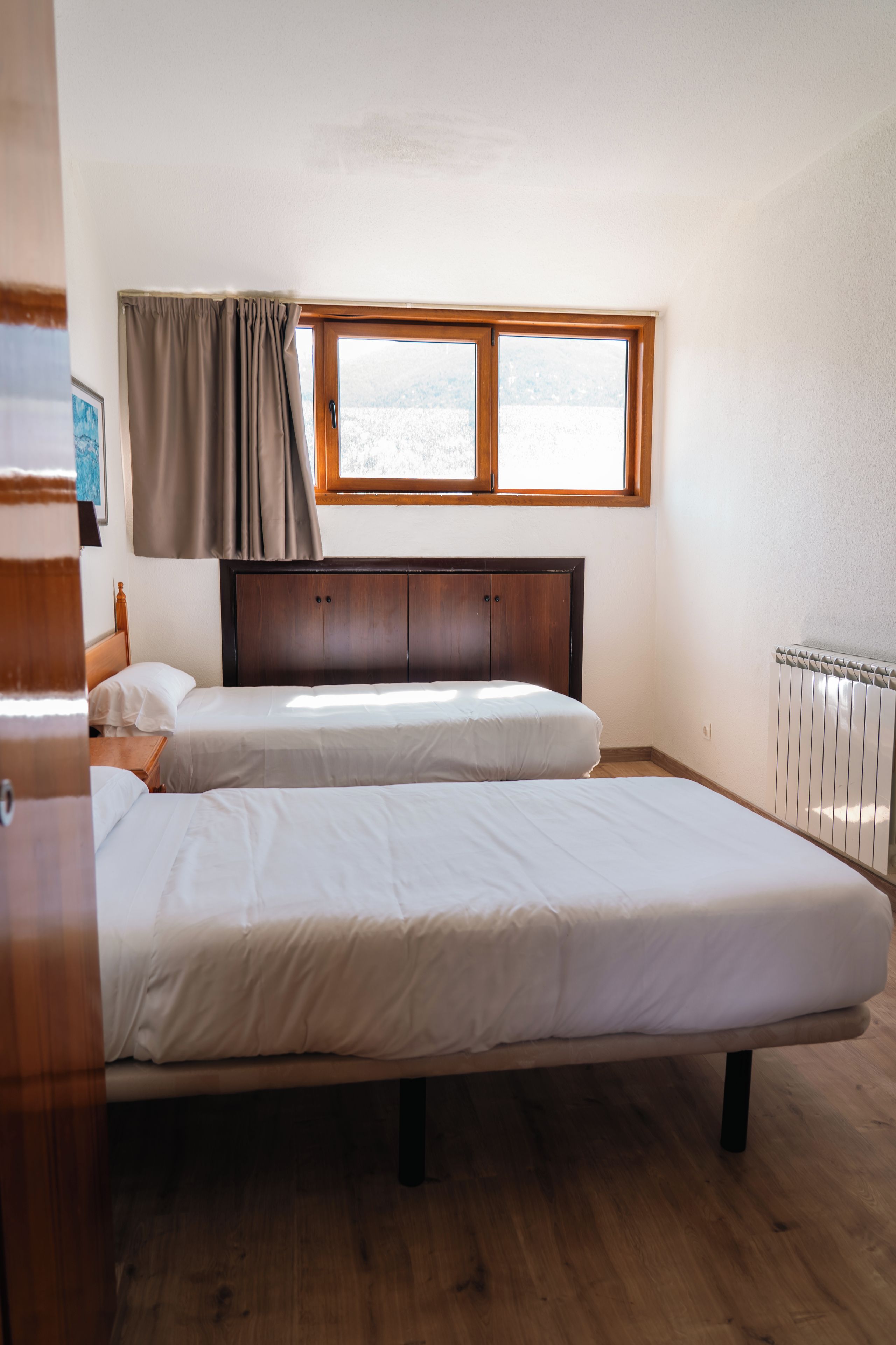 A room with two single beds, a wooden headboard, a window with curtains, and a radiator.