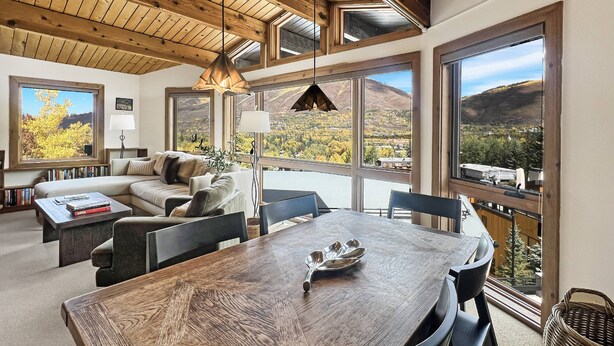 Een ruime woonkamer met houten plafond, grote ramen met uitzicht op bergen, een comfortabele bank en een eettafel met stoelen.