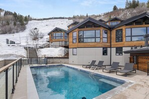 A modern building with a swimming pool, outdoor seating, and a snowy hillside in the background.