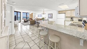 A spacious kitchen with a large island, bar stools, and a view of the ocean through the open door. - Phoenix VI 813 | Pools | Beachfront | Racquetball | My Beach Getaways (Orange Beach)