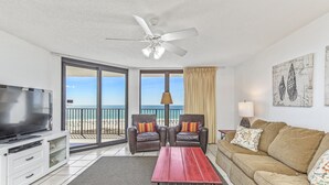A living room with a TV, a red table, a brown sofa, two armchairs, and a ceiling fan. - Phoenix VI 813 | Pools | Beachfront | Racquetball | My Beach Getaways (Orange Beach)