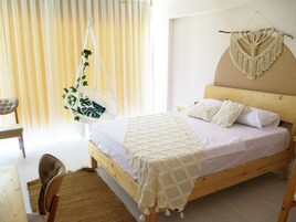 Un dormitorio con cama de madera, ropa de cama blanca, un tapiz de macramé en la pared, una silla colgante con plantas y vistas a una ventana con cortinas.