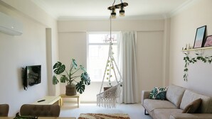 A living room with a sofa, a coffee table, a hanging chair, a TV mounted on the wall, and a large window with curtains. - Santa Center Pansiyon  (Demre)