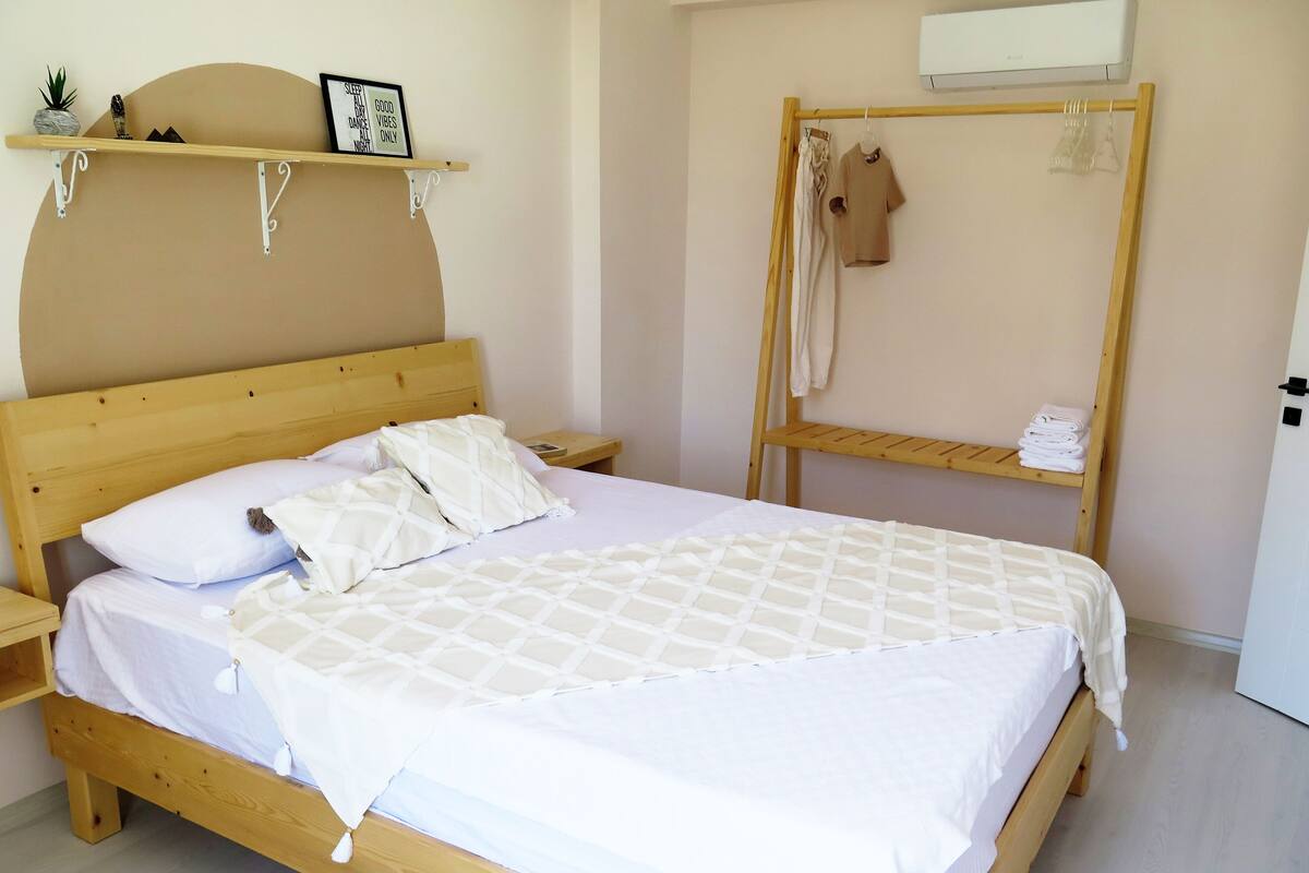 A wooden bed with white bedding, a bedside table, a shelf with decorative items, a bunk bed with a towel rack, and an air conditioning unit.