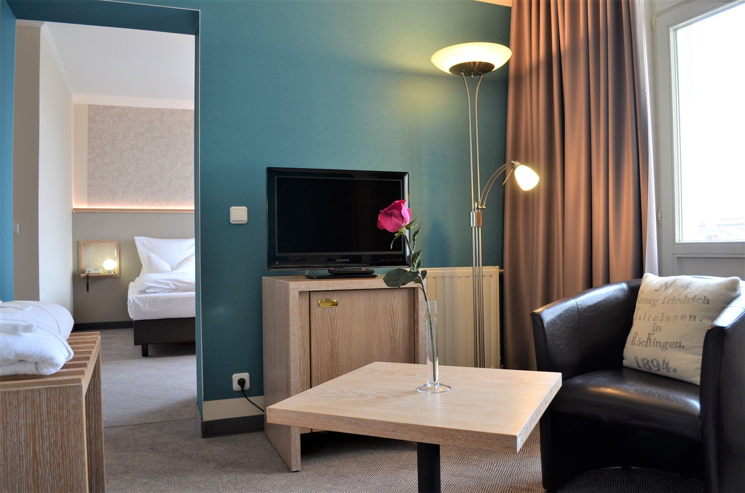 A hotel room with a bed, a television, a table with a vase, a lamp, and a sofa.