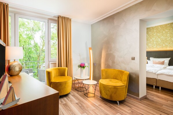 A hotel room with a wooden floor, two yellow armchairs, a small round table, a bed with white bedding, and a view of trees through large windows.