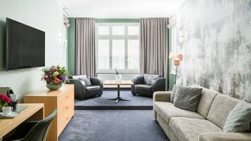 Ein modernes Hotelzimmer mit einem Flachbildfernseher, einem Holztisch mit Blumenvase, zwei Ledersofas, einem kleinen Tisch und einem großen Fenster mit Vorhängen.
