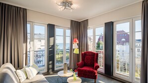 Una habitación con sofá, un sillón rojo, dos mesitas y ventanas amplias con vistas a edificios y una masa de agua.