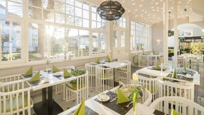 Un comedor luminoso con mobiliario blanco, ventanas amplias y una lámpara colgante central.