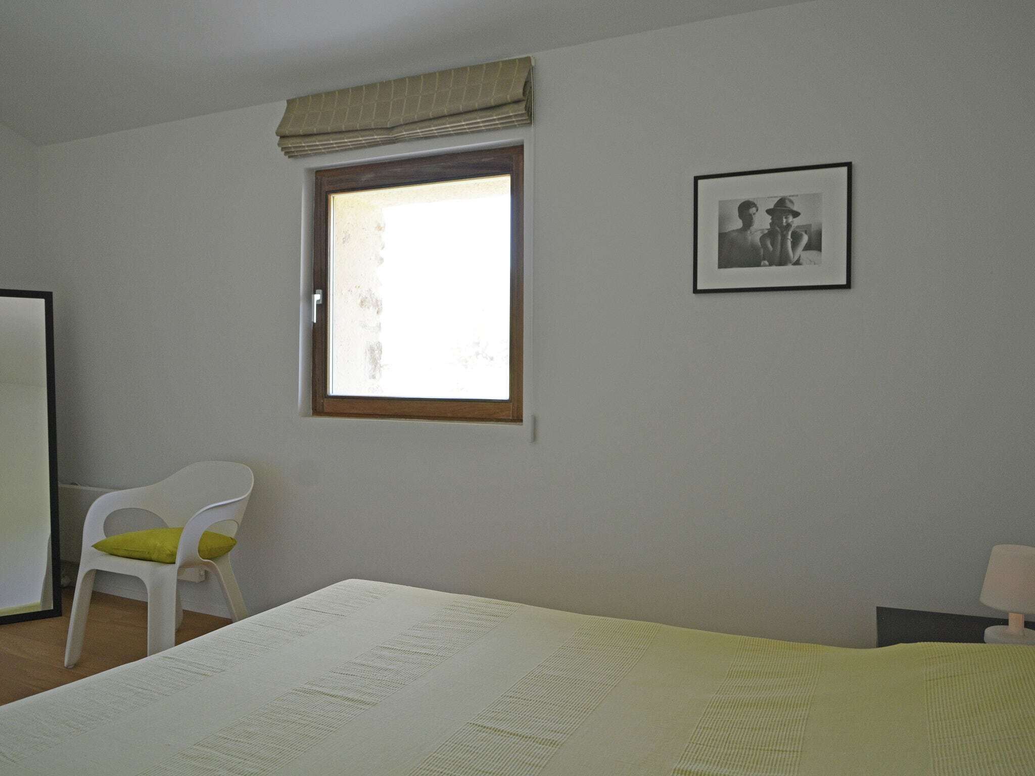 A bedroom with a bed, a window with a curtain, a white chair, and a framed picture on the wall.