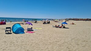 Una playa arenosa con tiendas de campaña y sombrillas, gente tomando el sol, y un cielo azul despejado.