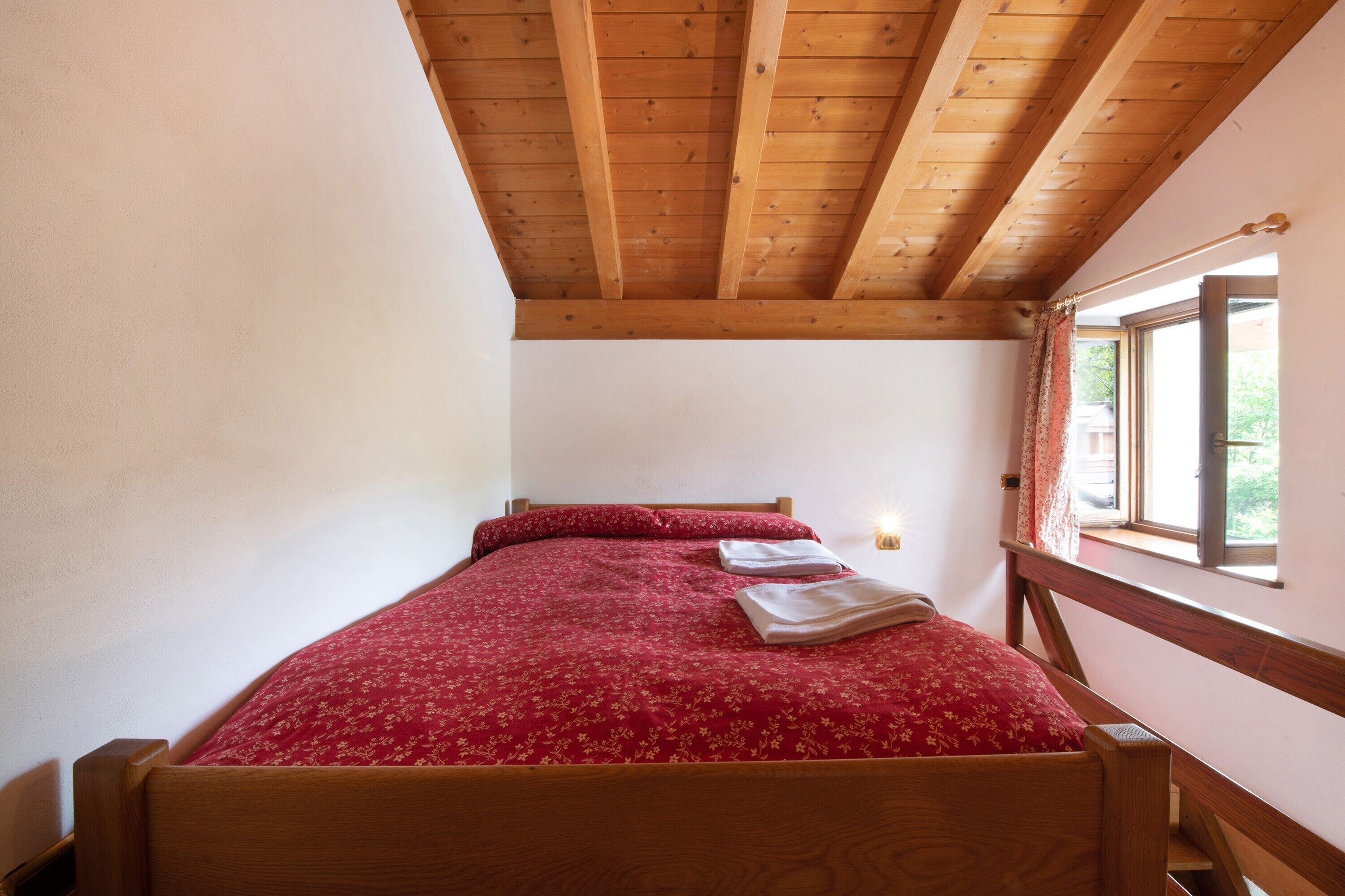 A single bed with a red floral bedspread, wooden headboard, and a window with curtains.