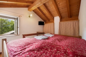 Ein Schlafzimmer mit Holzboden, einem Bett mit floralem Überwurf, einem Fenster mit Blick auf Bäume und einem an der Wand befestigten Fernseher.