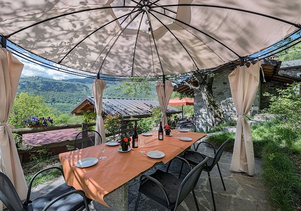 Ein Essbereich im Freien mit Tisch für zwei Personen unter einem großen Sonnenschirm mit Blick auf eine gebirgige Landschaft.