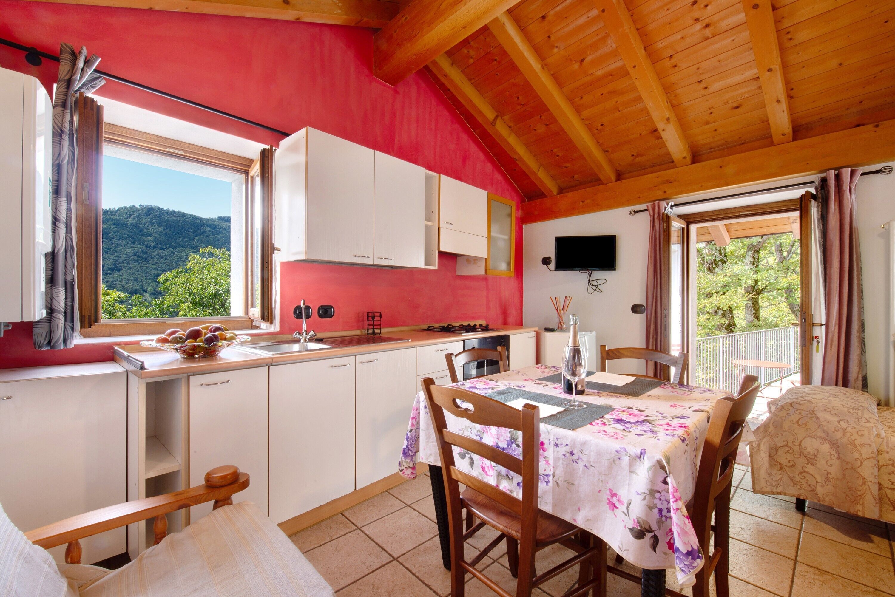 A kitchen with white cabinets, a dining table with floral tablecloth, wooden ceiling beams, and a view of greenery outside.