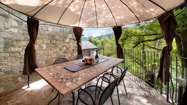 Una mesa con mantel de mosaico, un cuenco de fruta y una botella, bajo un paraguas grande en un balcón con paredes de piedra y vistas a zonas verdes.