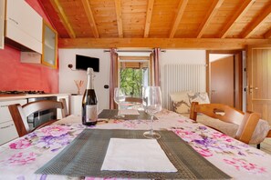 Un plafond en bois, une table à manger recouverte d’une nappe à motifs floraux, des chaises, une bouteille de vin et des verres à vin.
