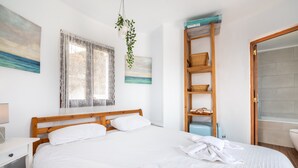 Una cama bien tendida con sábanas blancas, cabecera de madera, ventana con cortinas traslúcidas, una planta en la pared y un estante de madera con cestas.