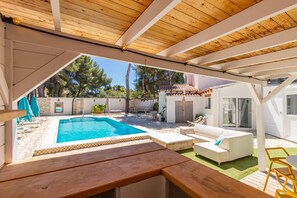 Ein überdachter Außenbereich mit Pool, weißen Möbeln und einem Holztisch.