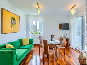 Uma sala de estar moderna com um sofá verde, mesa de jantar de madeira e cadeiras. Há uma janela com cortinas, uma TV de tela plana e um quadro na parede.