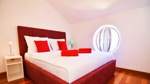 Quarto com cama estofada vermelha, roupa de cama branca e janela redonda com cortinas.
